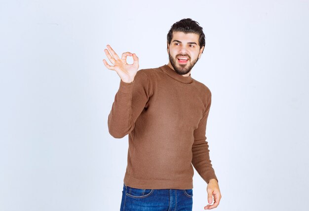 Hombre joven de pie en suéter marrón mostrando gesto bien. Foto de alta calidad