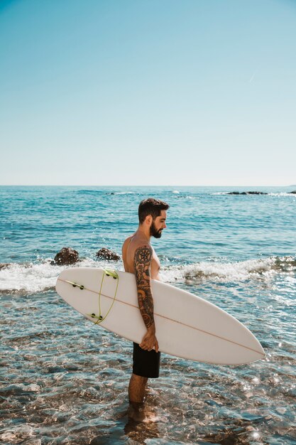 Hombre joven de pie con tabla de surf en agua azul