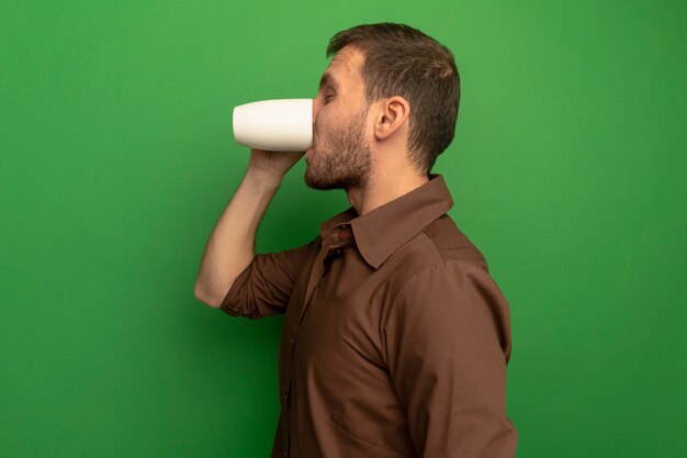 Hombre joven de pie en la vista de perfil bebiendo una taza de té con los ojos cerrados aislado en la pared verde