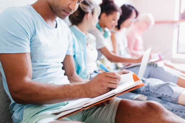 Hombre joven con piel morena en camisa azul escribiendo una conferencia en un cuaderno sentado al lado de compañeros de la universidad. Retrato interior de estudiantes que estudian juntos en la biblioteca de la universidad.