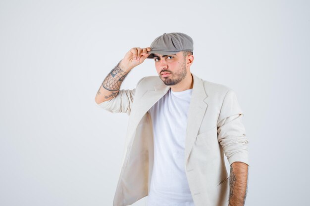 Hombre joven poniendo la mano en la gorra en camiseta blanca, chaqueta y gorra gris y mirando serio. vista frontal.