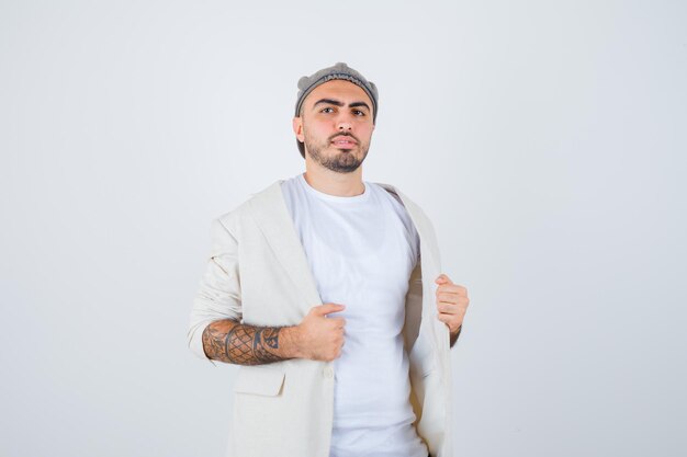 Hombre joven poniendo las manos en la chaqueta y tratando de quitarse la chaqueta con camiseta blanca, chaqueta y gorra gris y mirando serio. vista frontal.