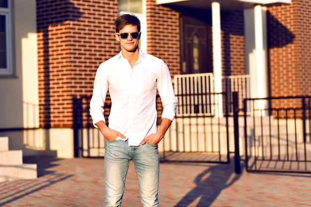 Hombre joven posando en la calle, look casual elegante, colores cálidos y soleados, joven empresario caminando solo, gafas de sol de moda, camisa blanca.