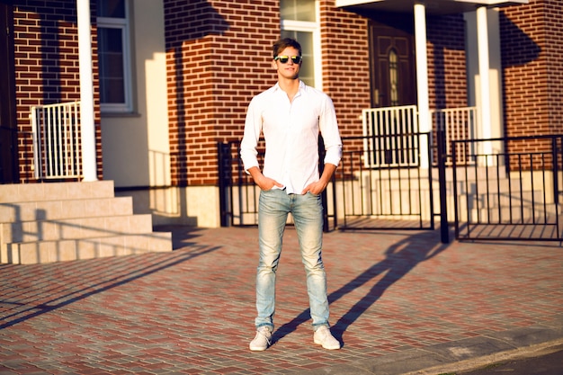 Hombre joven posando en la calle, look casual elegante, colores cálidos y soleados, joven empresario caminando solo, gafas de sol de moda, camisa blanca.