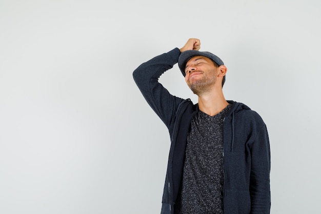 Hombre joven posando con la mano en la cabeza en camiseta, chaqueta, gorra y buscando pacífica.