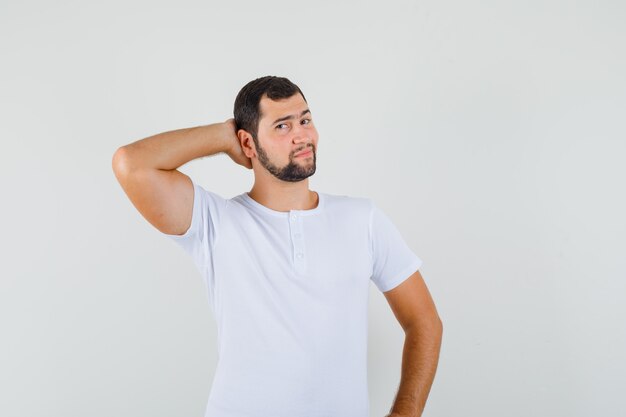 Hombre joven posando con la mano en el cuello en camiseta blanca y luciendo impresionante. vista frontal.