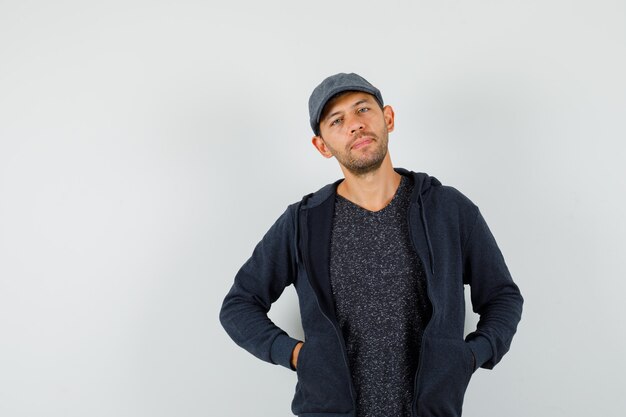 Hombre joven posando con las manos en los bolsillos en camiseta, chaqueta, gorra y elegante.