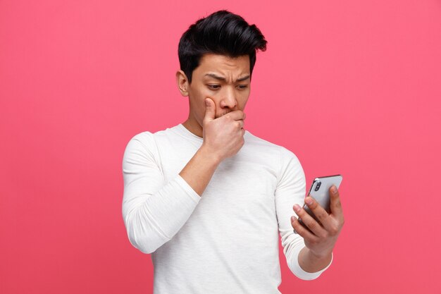 Hombre joven preocupado sosteniendo y mirando el teléfono móvil manteniendo la mano en la boca