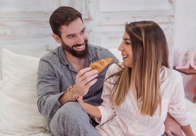 Hombre joven que alimenta a la mujer con el croissant en cama