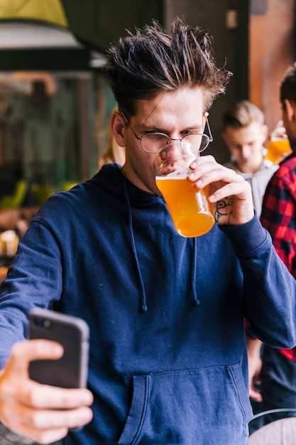 Foto gratuita hombre joven que bebe el vidrio de cerveza que toma el selfie en smartphone