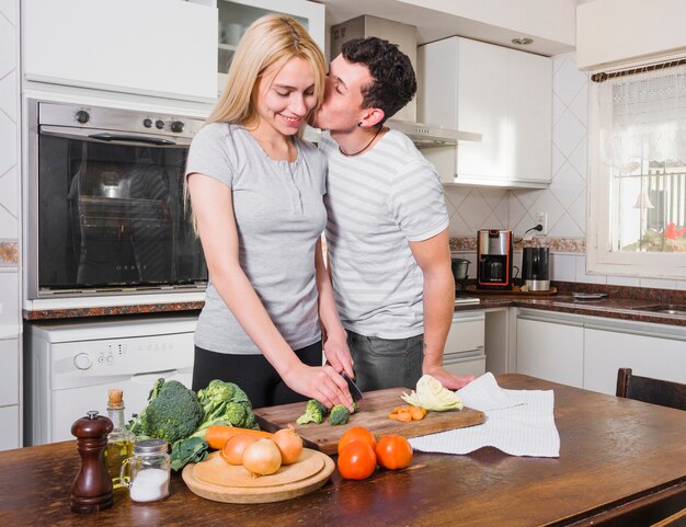 Hombre joven que besa a su esposa que corta la verdura en tajadera