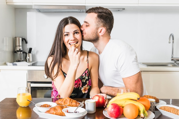 Foto gratuita hombre joven que besa a su novia comiendo galletas con frutas y croissant en la mesa en la cocina