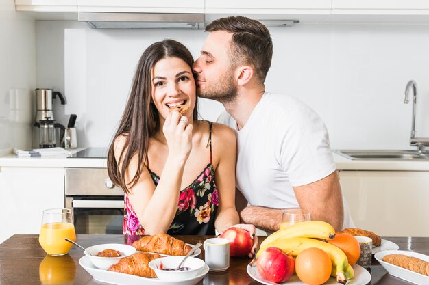 Hombre joven que besa a su novia comiendo galletas con frutas y croissant en la mesa en la cocina