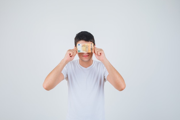 Hombre joven que cubre los ojos con billetes en euros en camiseta y mirando alegre. vista frontal.