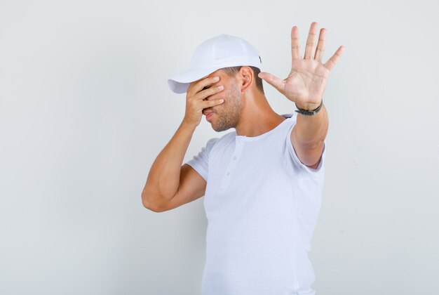 Hombre joven que cubre los ojos y hace el gesto de parada en camiseta blanca, gorra, vista frontal.