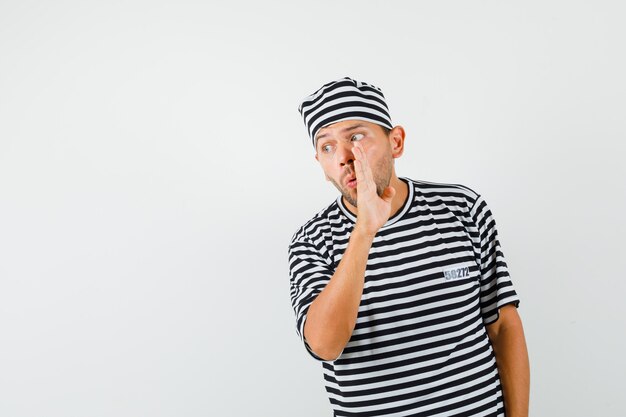 Hombre joven que dice el secreto detrás de la mano en camiseta rayada, sombrero.