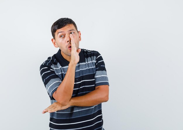 Hombre joven que dice el secreto detrás de la mano en la vista frontal de la camiseta.