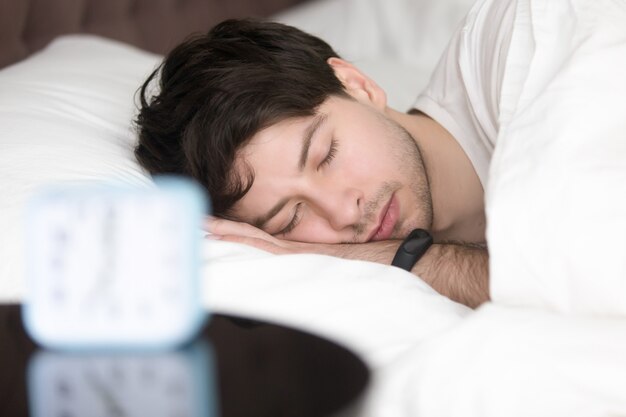 Hombre joven que duerme al lado del reloj de alarma que lleva pulsera inteligente