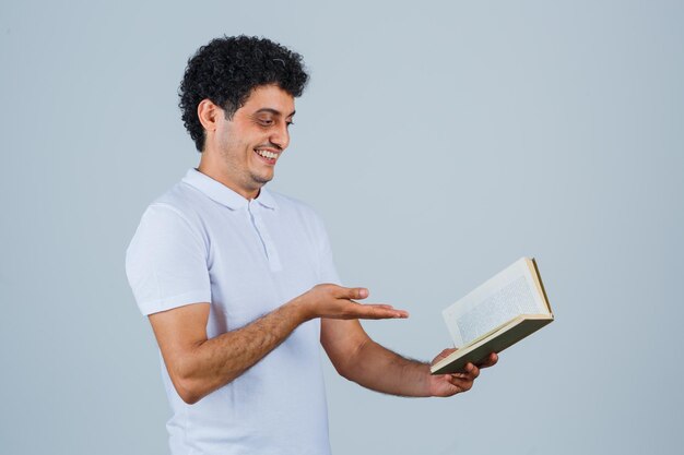 Hombre joven que estira la mano hacia el libro en camiseta blanca y jeans y parece feliz. vista frontal.