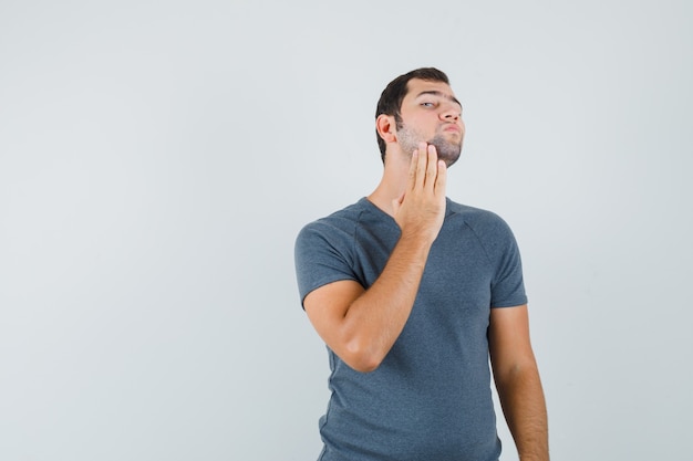 Hombre joven que examina la piel de la cara tocando su barba en una camiseta gris y luciendo guapo