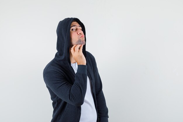 Hombre joven que examina la piel tocando su barba en la chaqueta, camiseta y luciendo guapo, vista frontal.