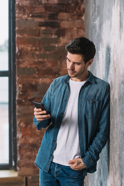 Hombre joven que se inclina delante de la pared usando el teléfono celular