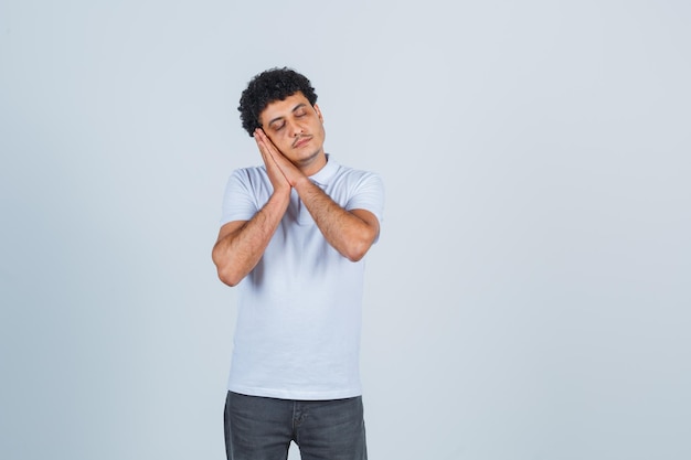 Hombre joven que se inclina la mejilla en las manos, fingiendo que quiere dormir con una camiseta blanca y pantalones vaqueros y que parece soñoliento, vista frontal.