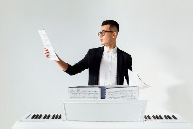 Hombre joven que lee la hoja musical que está parada detrás del piano contra el fondo blanco