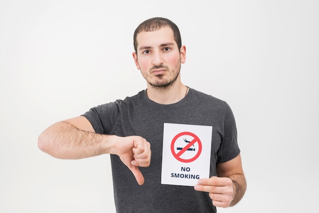 Un hombre joven que lleva a cabo la muestra de no fumadores que muestra los pulgares hacia abajo contra el fondo blanco