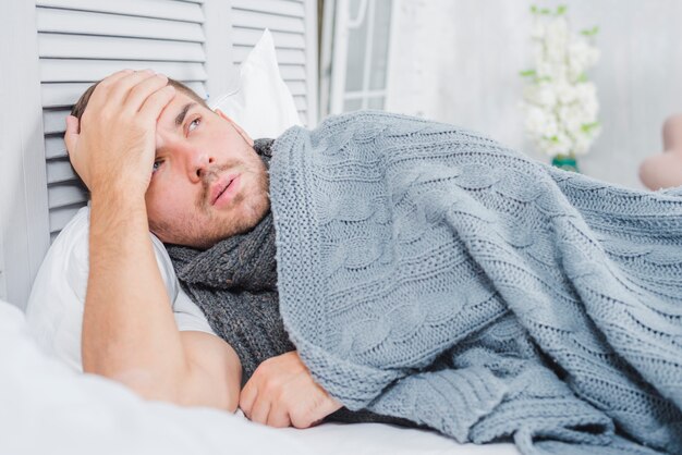 Hombre joven que miente en cama con dolor de cabeza y fiebre que toca su frente