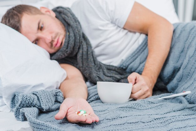 Hombre joven que miente en la cama que sostiene píldoras y la taza disponibles