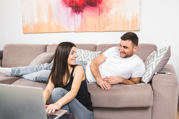 Hombre joven que miente en el sofá que mira a su novia que usa el ordenador portátil