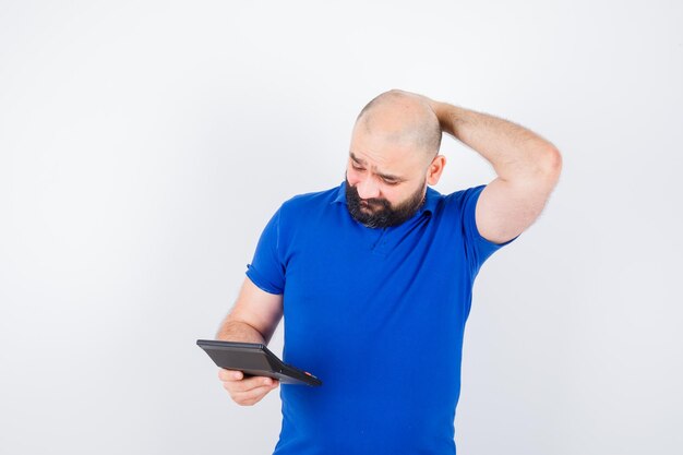 Hombre joven que mira la calculadora mientras sostiene la mano en la cabeza con la camisa azul y parece perdido. vista frontal.