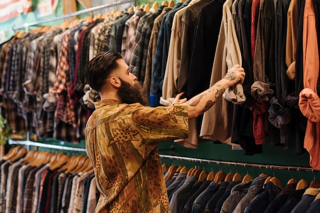 Hombre joven que mira la camisa que cuelga en el carril dentro de la tienda de ropa