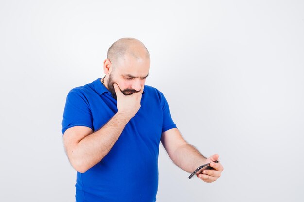 Hombre joven que mira el teléfono mientras piensa en camisa azul y parece pensativo. vista frontal.