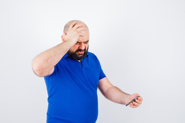 Hombre joven que mira el teléfono mientras sostiene la mano en la cabeza con la camisa azul y parece agresivo, vista frontal.