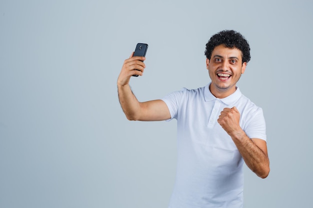 Hombre joven que mira el teléfono móvil en la camiseta blanca y que parece feliz. vista frontal.