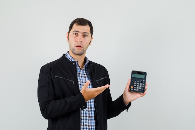 Hombre joven que muestra la calculadora en camisa, chaqueta y mirando perplejo, vista frontal.