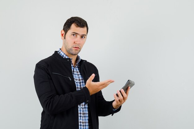 Hombre joven que muestra la calculadora en camisa, chaqueta y mirando sensible, vista frontal.