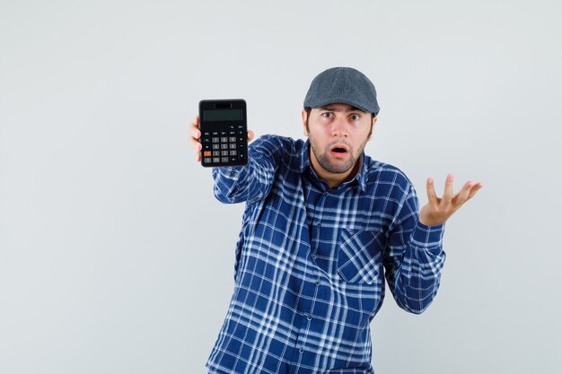 Hombre joven que muestra la calculadora en camisa, gorra y mirando perplejo. vista frontal.