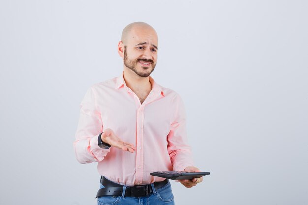 Hombre joven que muestra la calculadora en camisa rosa, jeans, vista frontal.