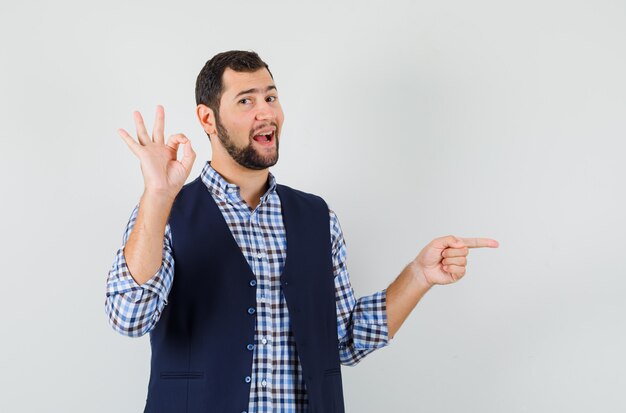 Hombre joven que muestra un gesto bien, apuntando hacia el lado en camisa, chaleco y mirando feliz