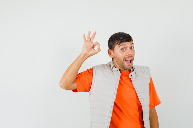 Hombre joven que muestra gesto bien en camiseta, chaqueta y mirando feliz.