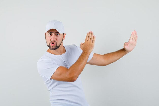 Hombre joven que muestra el gesto de chuleta de karate en camiseta blanca, gorra y mirando flexible. vista frontal.