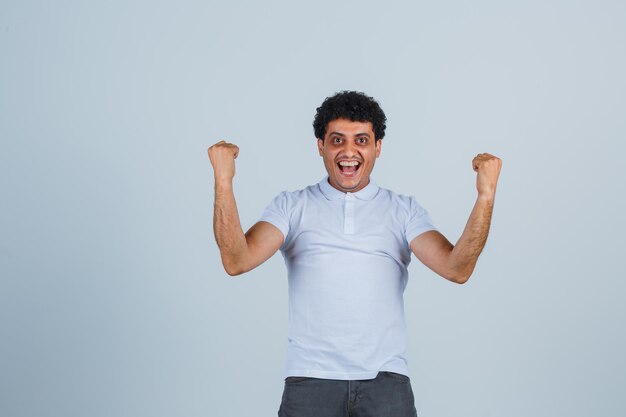 Hombre joven que muestra el gesto del ganador en la camiseta blanca y los pantalones vaqueros y parece feliz. vista frontal.