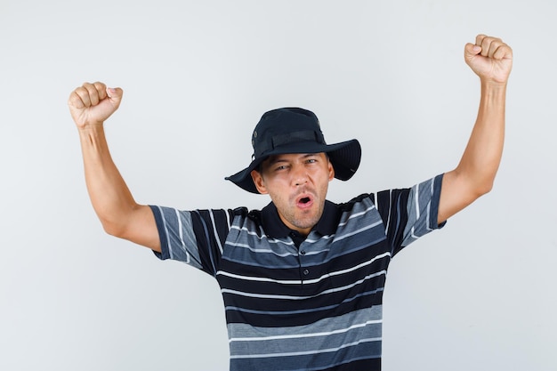 Hombre joven que muestra el gesto del ganador en camiseta, sombrero y que parece feliz. vista frontal.