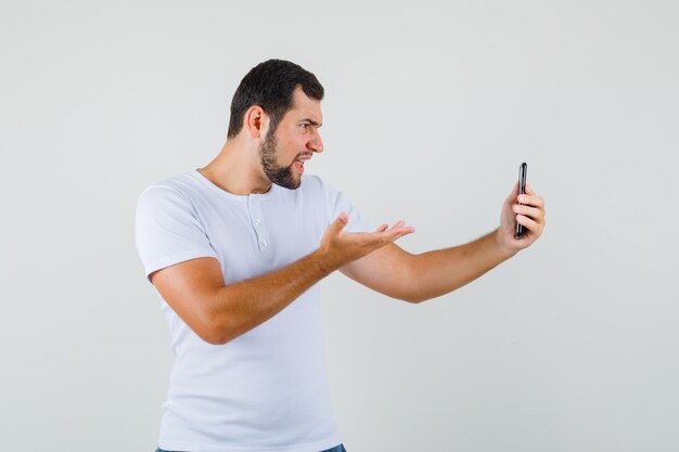 Hombre joven que muestra un gesto de interrogación mientras hace una videollamada en camiseta blanca y parece enojado.