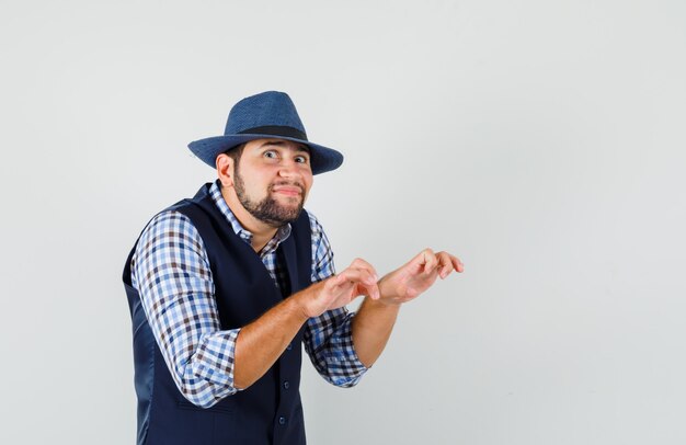 Foto gratuita hombre joven que muestra el gesto de mecanografía con los dedos apretados en camisa, chaleco, sombrero y mirando divertido.