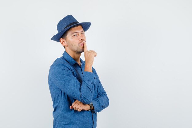 Hombre joven que muestra gesto de silencio en camisa azul, sombrero y mirando con cuidado. vista frontal.