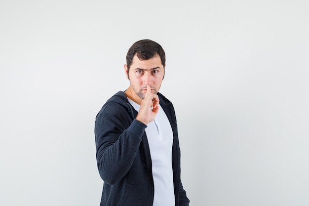 Hombre joven que muestra gesto de silencio en camiseta blanca y sudadera con capucha negra con cremallera frontal y aspecto serio. vista frontal.